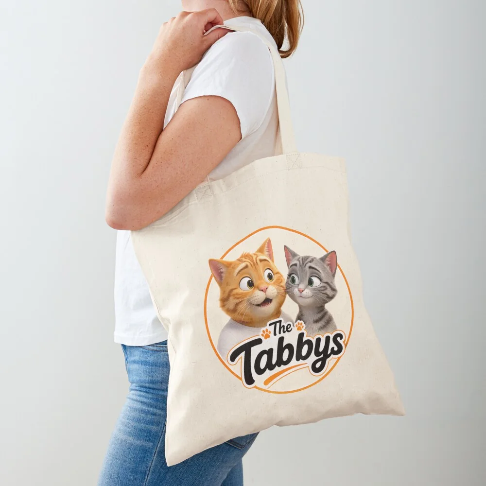 Person carrying a cotton tote bag featuring The Tabbys logo with George and Maggie the tabby cats.